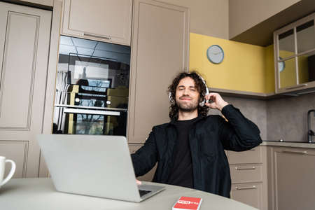 KYIV, UKRAINE - APRIL 25, 2020: smiling freelance in wireless headphones looking at camera near laptop and smartphone with Youtube app on screenのeditorial素材