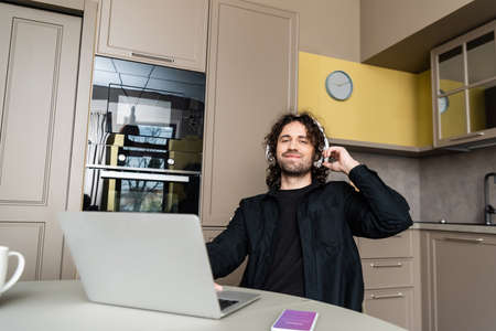 KYIV, UKRAINE - APRIL 25, 2020: Selective focus of smiling freelancer in headphones using laptop near smartphone with instagram app on kitchen tableのeditorial素材