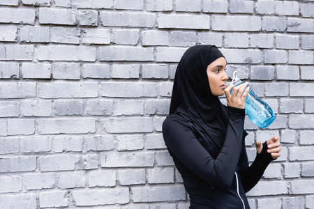 young muslim sportswoman in hijab holding sports bottle while drinking water outside