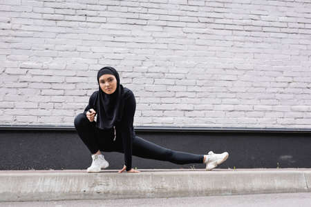 arabian sportswoman in hijab stretching near brick wall
