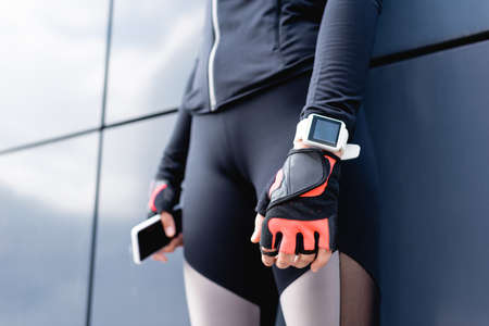 cropped view of young sportswoman with fitness tracker on hand standing and holding smartphone