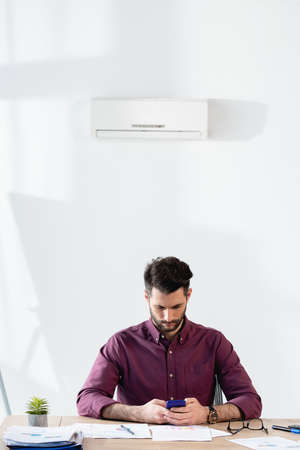 young businessman using smartphone while sitting at workplace under air conditioner