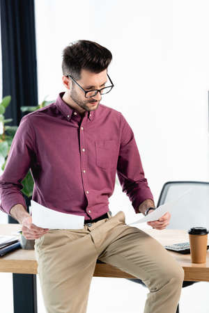 attentive businessman holding documents while sitting on desk near coffee to go