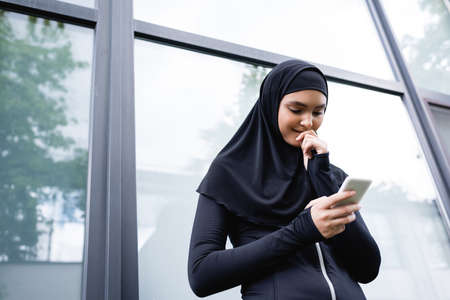 low angle view of cheerful muslim woman looking at smartphone