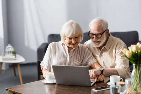 Selective focus of smiling elderly couple using laptop near coffee cups on table