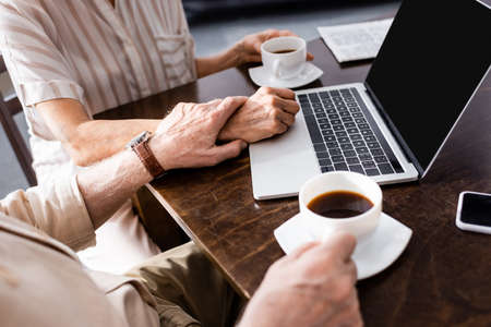 Cropped view of elderly man touching hand of wife near cups of coffee and digital devices on tableの写真素材