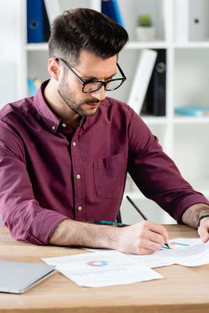 attentive businessman in eyeglasses writing on paper while sitting at workplaceの写真素材