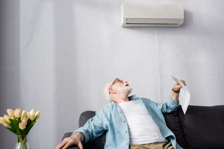 Selective focus of elderly man holding newspaper and looking at air conditioner at homeの写真素材