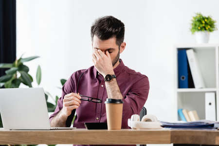 exhausted businessman holding eyeglasses and touching eyes while sitting near laptop