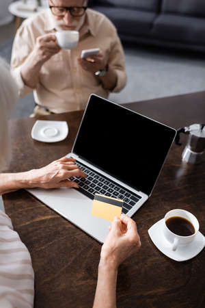 Selective focus of senor woman using laptop and credit card near husband with coffee and smartphone...