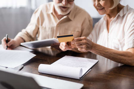 Selective focus of senior woman holding credit card while husband writing on papers near gadgets on...