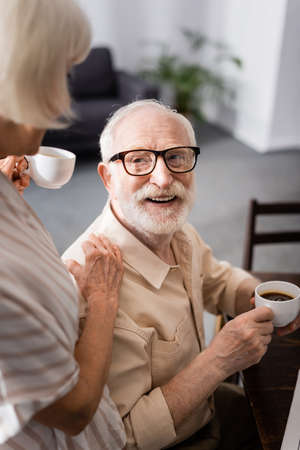 Selective focus of senior man holding cup of coffee near wife at homeの写真素材