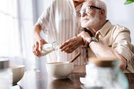Selective focus of man hugging senor wife pouring milk in bowl at table
