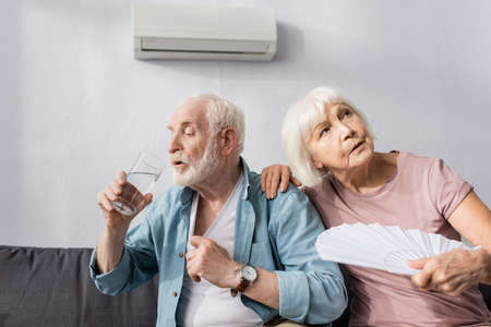 Elderly woman holding fan near husband drinking water on couch at home