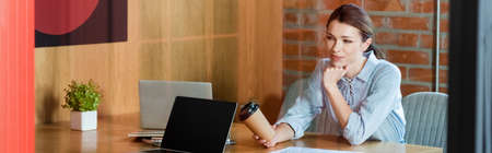 panoramic concept of businesswoman holding paper cup near laptops