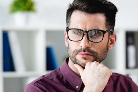 confident, pensive businessman in eyeglasses touching chin while looking away