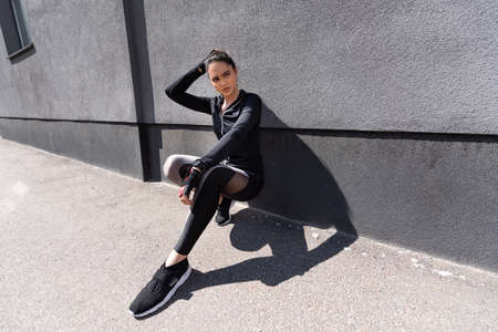 beautiful sportswoman touching hair while sitting near concrete wallの写真素材