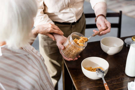 Selective focus of senior woman touching hands of husband pouring cereals in bowl