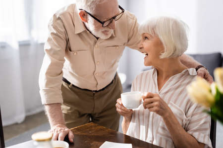 Selective focus of senior man hugging positive wife with cup of coffee in kitchenの写真素材