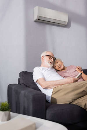 Selective focus of smiling elderly man holding remote controller of air conditioner near wife on couch at homeの写真素材