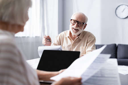 Selective focus of smiling man looking at wife with papers near laptop at homeの写真素材