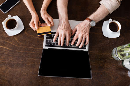 Top view of senior couple using laptop and credit card near smartphone and coffee cups on tableの写真素材