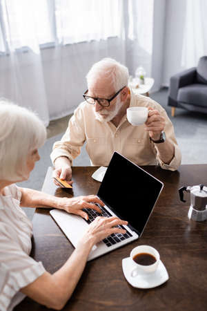 Selective focus of senior man holding credit card and cup of coffee near wife using laptop at home