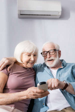 Selective focus of smiling senior man embracing wife and using remote controller of air conditioner at homeの写真素材