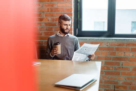 selective focus of bearded businessman holding coffee to go and reading newspaper near laptopの写真素材