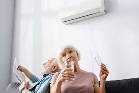 Selective focus of elderly woman holding glass of water and fan near husband suffering from heat at homeの写真素材