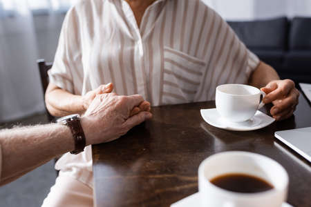 Cropped view of senor couple holding hands while drinking coffee at tableの写真素材