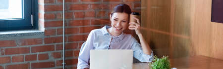 panoramic crop of happy businesswoman holding paper cup with coffee to go while looking at laptop