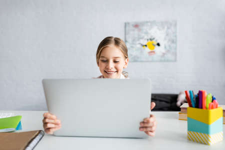 Selective focus of smiling child holding laptop near books on tableの写真素材