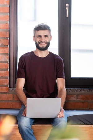 selective focus of cheerful businessman sitting on window bench and holding laptopの写真素材