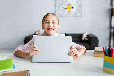 Selective focus of smiling kid holding laptop and looking at camera near stationeryの写真素材