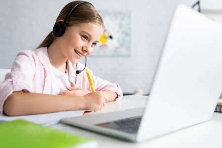 Selective focus of positive kid in headset writing on notebook and using laptop at home