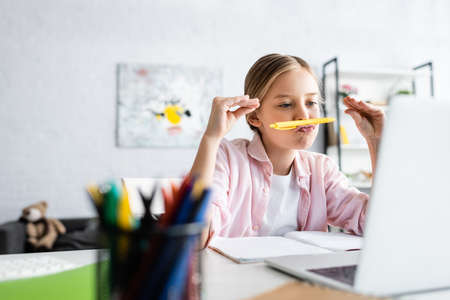 Selective focus of kid holding pen near mouth during webinar on laptop at home