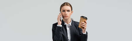 confused young businesswoman with coffee to go talking on smartphone isolated on gray, panoramic shotの写真素材