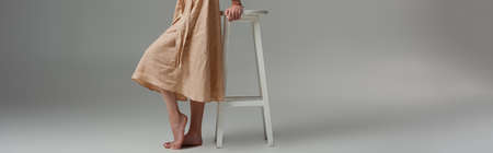 panoramic crop of barefoot woman standing near chair on gray