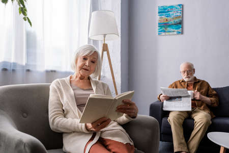attentive senior woman reading book in armchair near husband with newspaper on sofaの写真素材