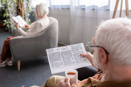selective focus of senior man holding cup of tea and reading newspaper while his wife reading book in armchairの写真素材