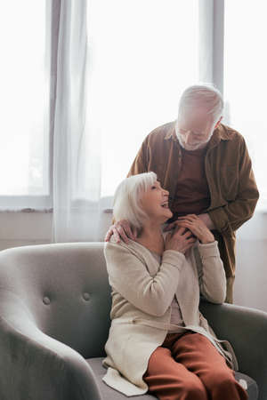senior man holding hand and touching shoulder of cheerful wife sitting in armchairの写真素材