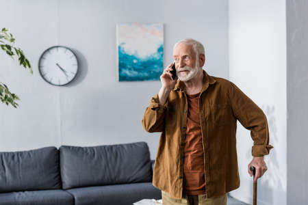 attentive senior man talking on smartphone while standing with walking stick