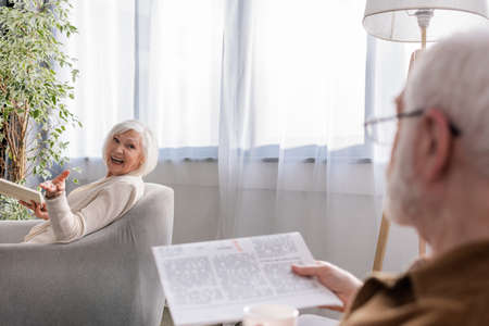 happy woman sitting in armchair with book while talking to husband with newspaper