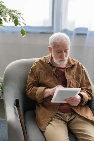 attentive senior man using digital tablet while sitting in armchair