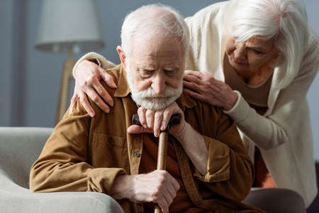 senior man, sick on dementia, sitting with walking stick, while wife looking at him and touching his shouldersの写真素材