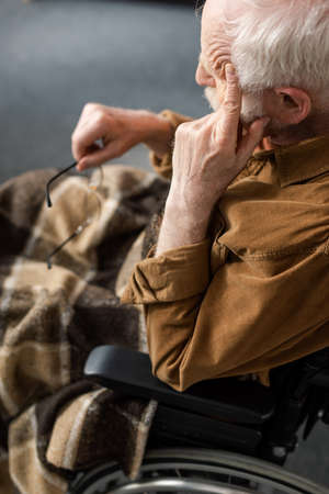 disabled senior man touching face and holding eyeglasses while sitting in wheelchairの写真素材