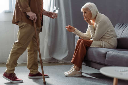 cropped view of senior man giving glass of water to wife feeling bad and sitting on sofaの写真素材