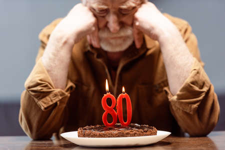 selective focus of lonely senior man leaning on hands while looking at birthday cake with number...