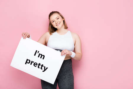 cheerful overweight girl holding placard with i am pretty inscription on pinkの写真素材